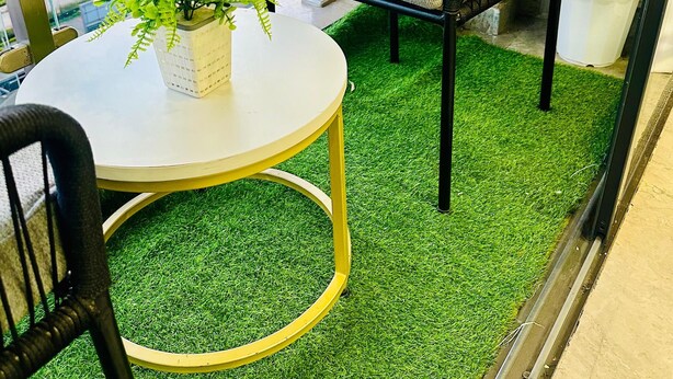 Een balkon met kunstgras, een ronde tafel, een zwarte rieten stoel met kussen, en een bloempot met plant.