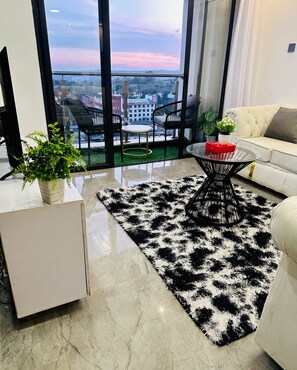 A modern living room with a large window, a white sofa, a black and white rug, and a small round table. - 1n2 bedroom w/gym 7 min walk to yaya and prestige mall, higher floor w/nice view (Nairobi)