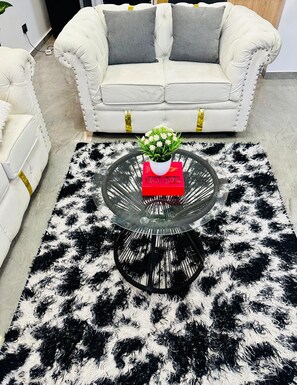 A living room with a white sofa, a glass coffee table with a red box, and a black and white furry rug. - 1n2 bedroom w/gym 7 min walk to yaya and prestige mall, higher floor w/nice view (Nairobi)