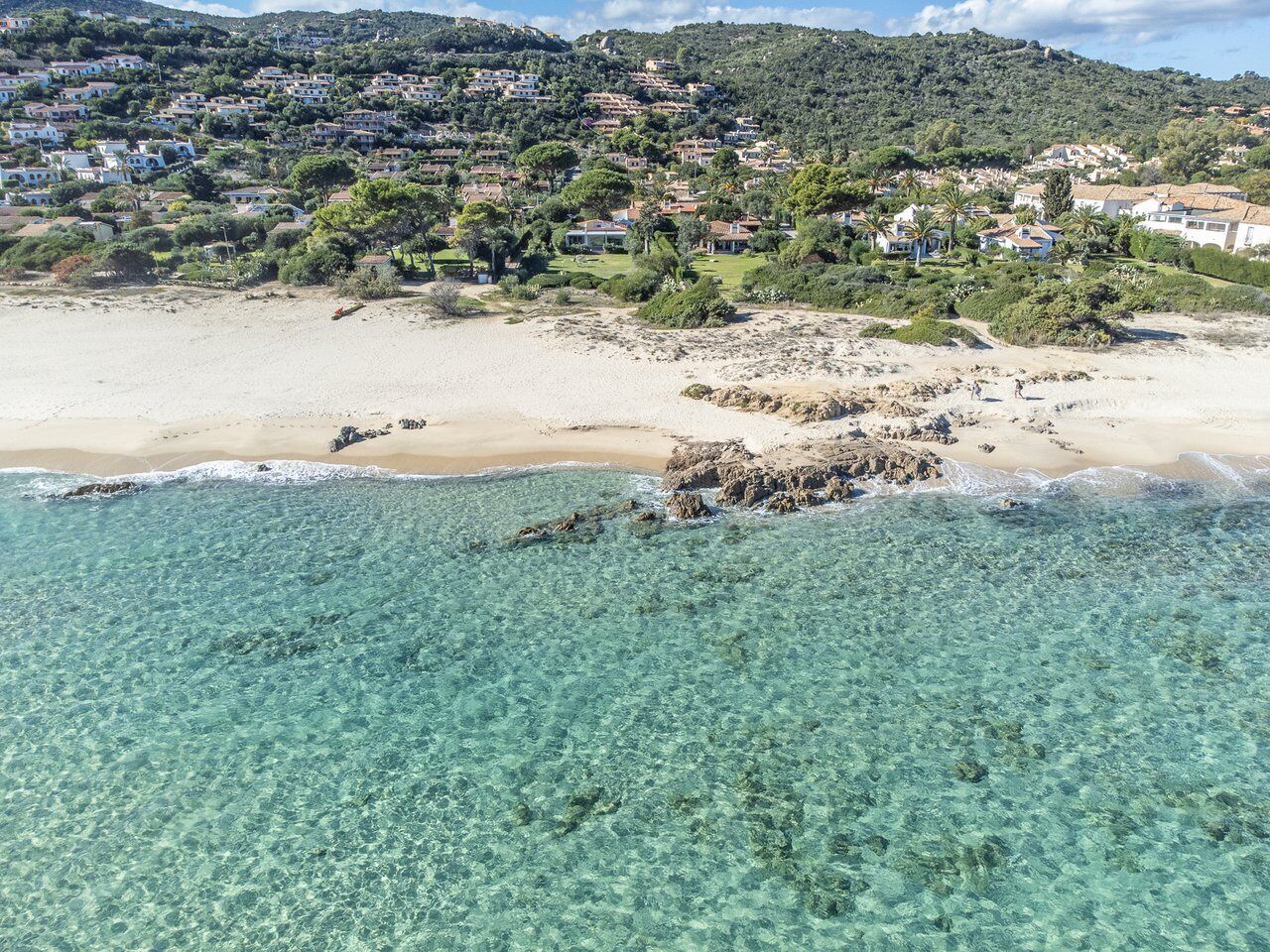 A coastal area with clear turquoise waters, sandy beach, and a hillside with residential buildings.