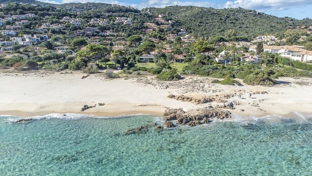 Zona costera con aguas turquesas cristalinas, playa de arena y un cerro con edificaciones residenciales.