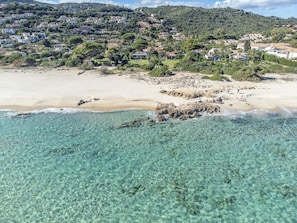 Kystområde med klart turkis vand, sandstrand og en bakke med boliger.