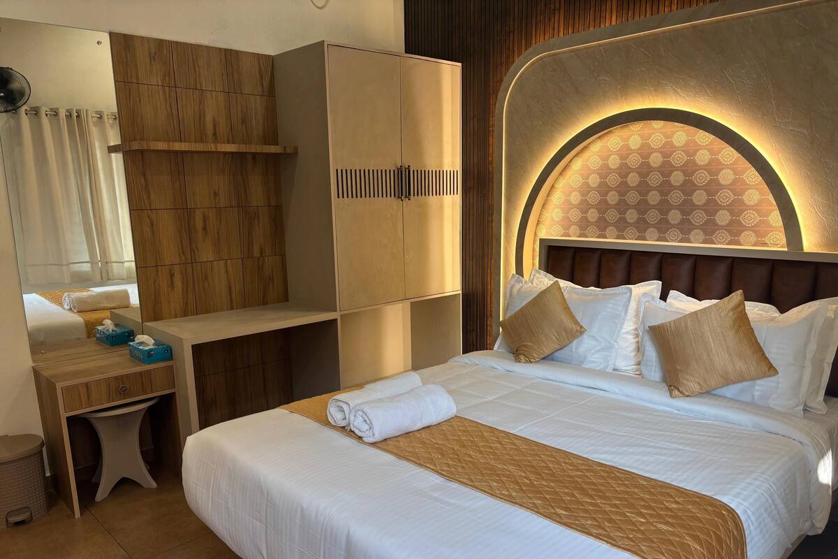 A hotel room with a large bed, a wooden wardrobe, a vanity with a mirror, and a patterned wall.