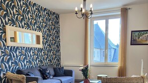 Un salon avec un canapé bleu, deux fauteuils en osier, une table basse, un lustre, une grande fenêtre avec des rideaux et un mur à motifs.