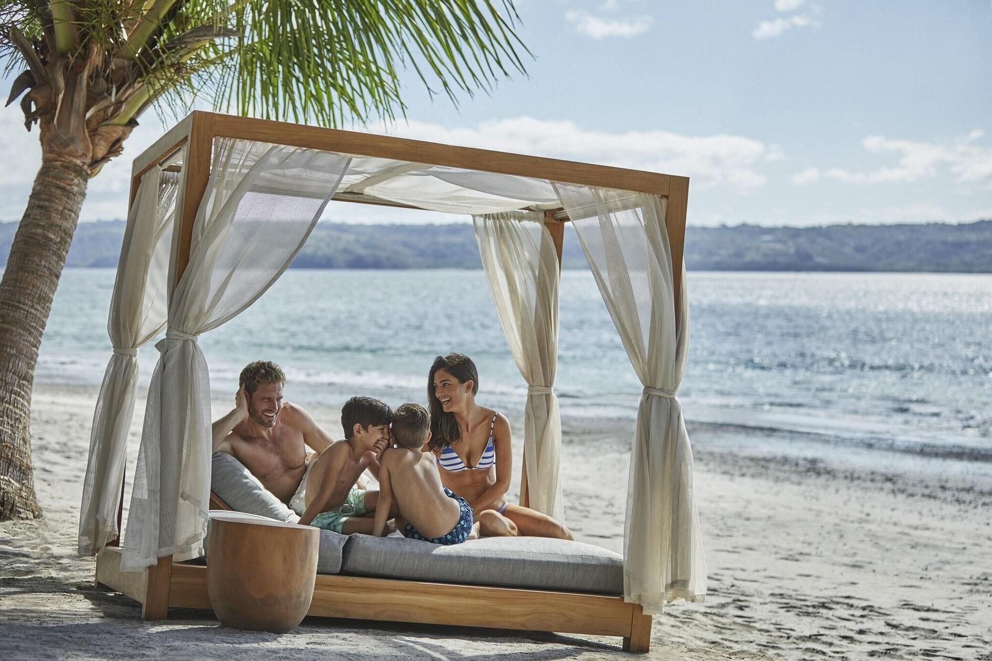 A beach cabana with a wooden frame and white curtains, a cushioned seating area, and a wooden bowl.