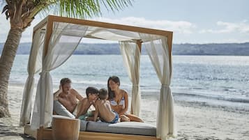 A beach cabana with a wooden frame and white curtains, a cushioned seating area, and a wooden bowl.