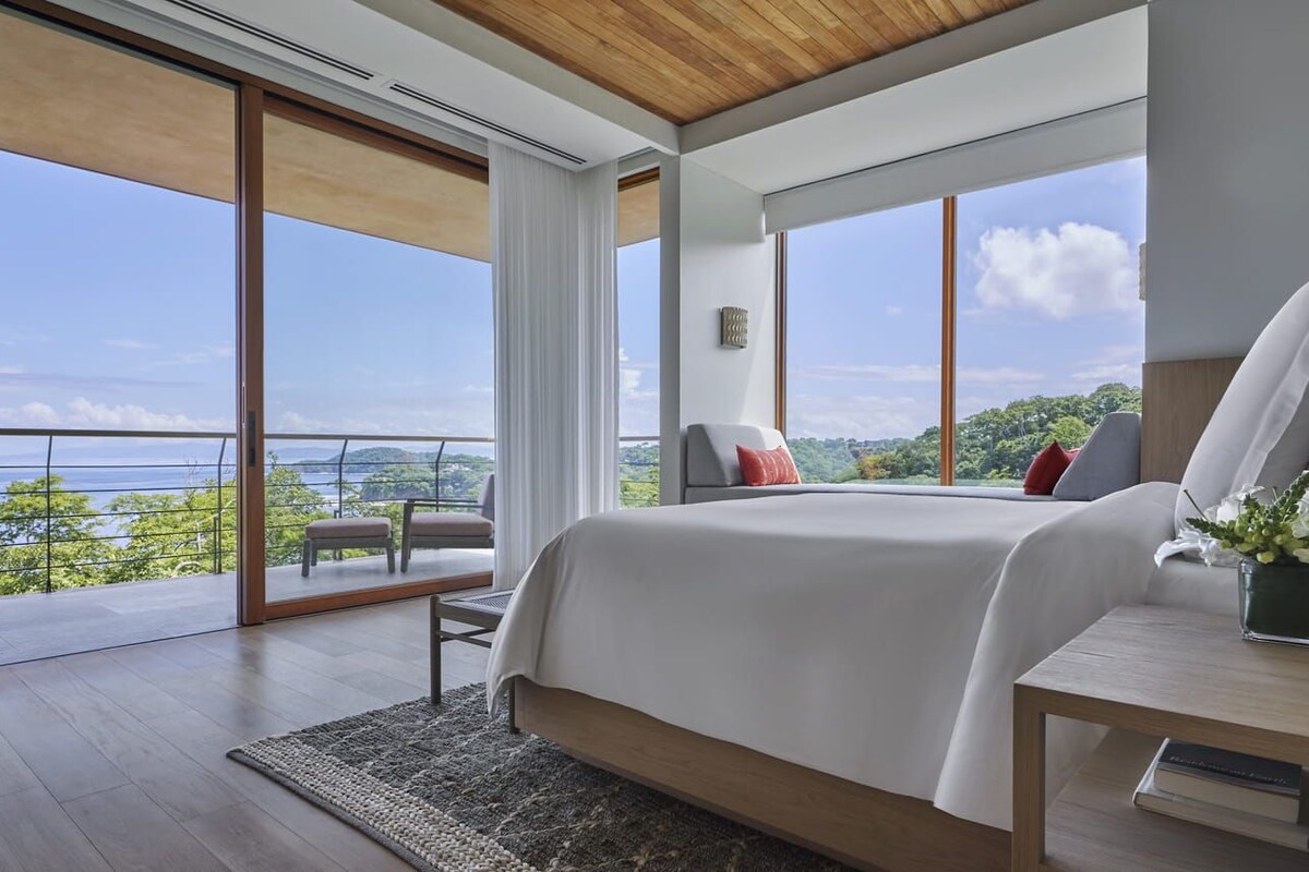 A bedroom with a large bed, a bedside table, a chair, and a view of the outdoors.