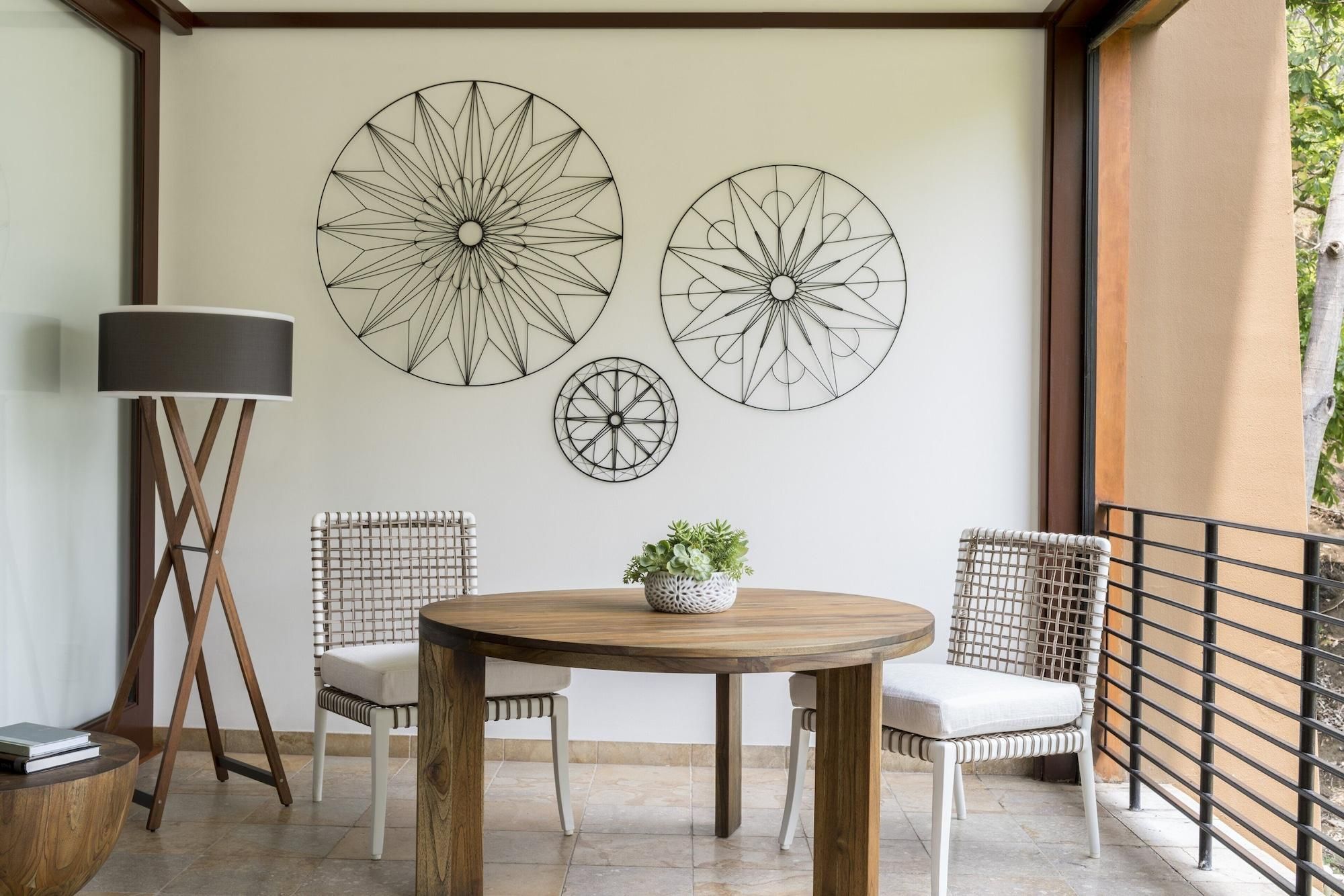 A round wooden table with two white cushioned chairs, a floor lamp, and three wall-mounted abstract art pieces.