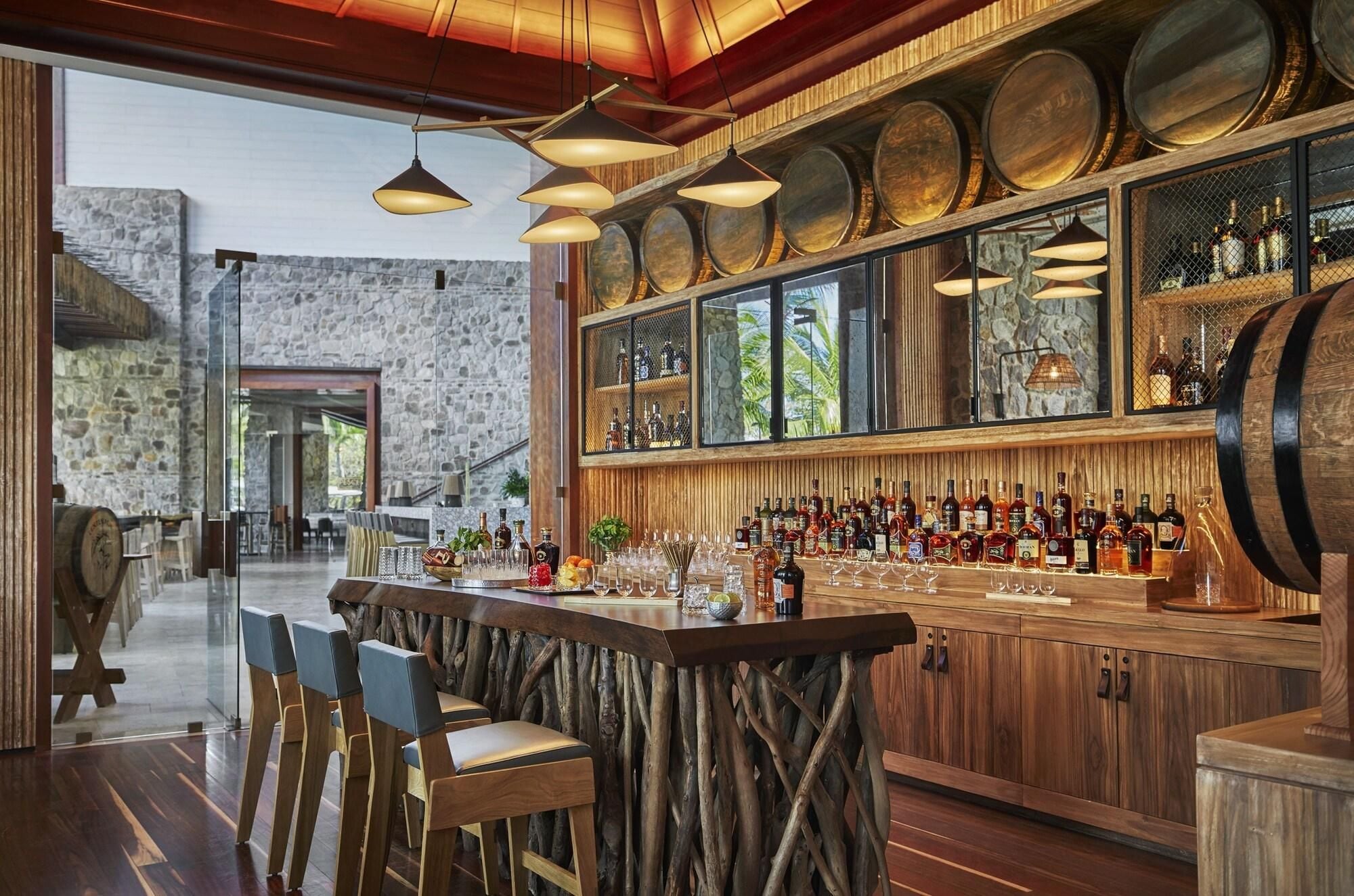 A bar area with wooden stools, a bar counter with bottles, and a stone wall in the background.