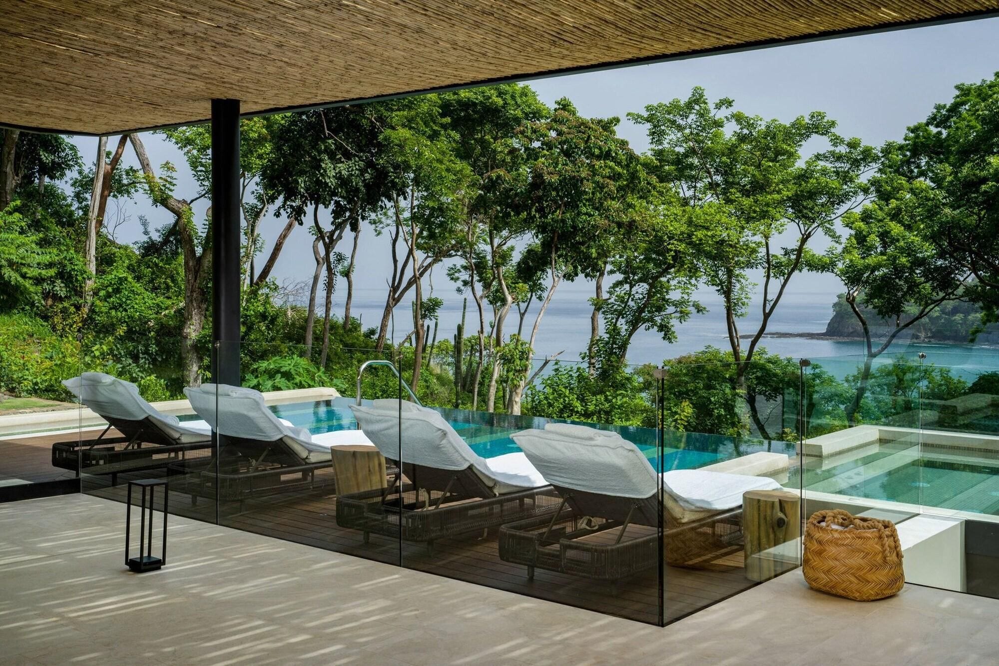 Un espacio lounge moderno al aire libre con paredes de vidrio, una piscina y vistas al mar.