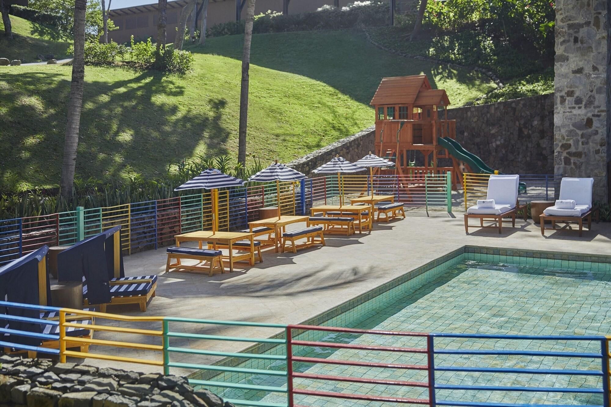 Zona de piscina con sillas de descanso, sombrillas y un pequeño edificio con techo rojo.