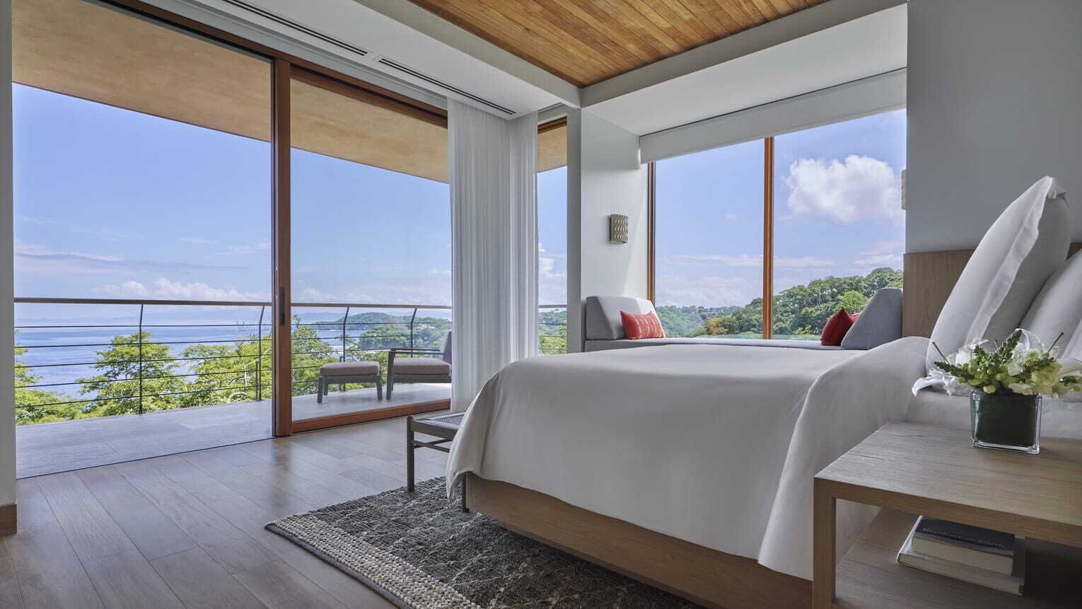 A bedroom with a large bed, a bedside table, a chair, and a view of the outdoors.