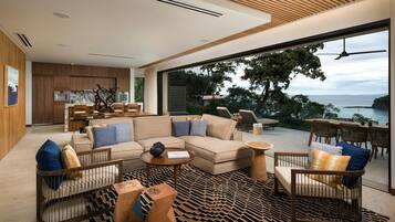A modern living room with a sofa, coffee table, and chairs, a kitchen in the background, and a large window with a view of the sea.