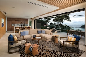 A modern living room with a sofa, coffee table, and chairs, a kitchen in the background, and a large window with a view of the sea. - Four Seasons Resort Costa Rica At Peninsula Papaga (Nacascolo)