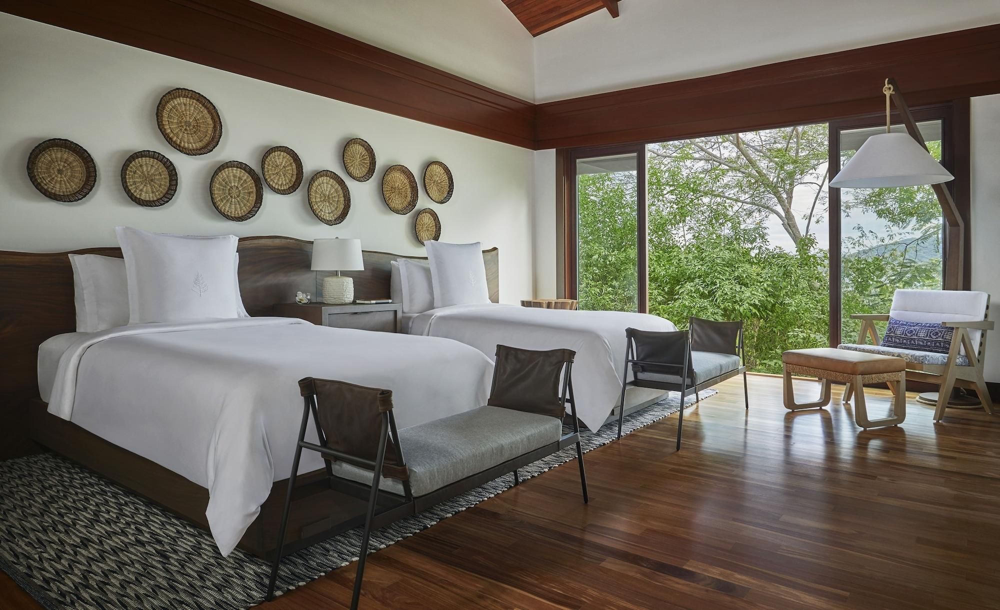 A hotel room with two beds, a sitting area with chairs, a lamp, and a view of greenery.