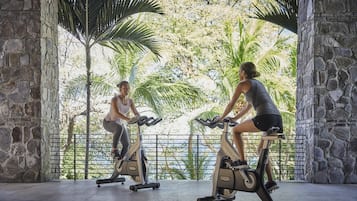 Two people exercising on stationary bikes in a room with stone walls and a view of greenery.