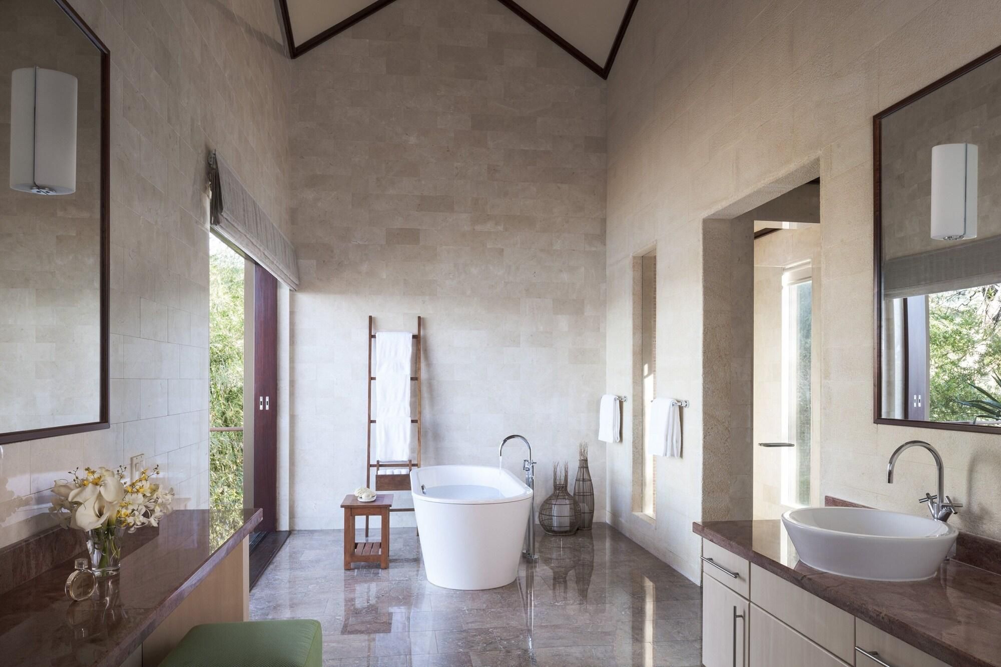 A modern bathroom with a freestanding bathtub, two sinks, a large mirror, and a view of greenery outside.