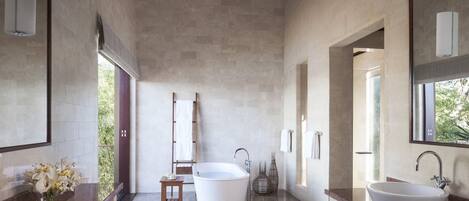 A modern bathroom with a freestanding bathtub, two sinks, a large mirror, and a view of greenery outside.