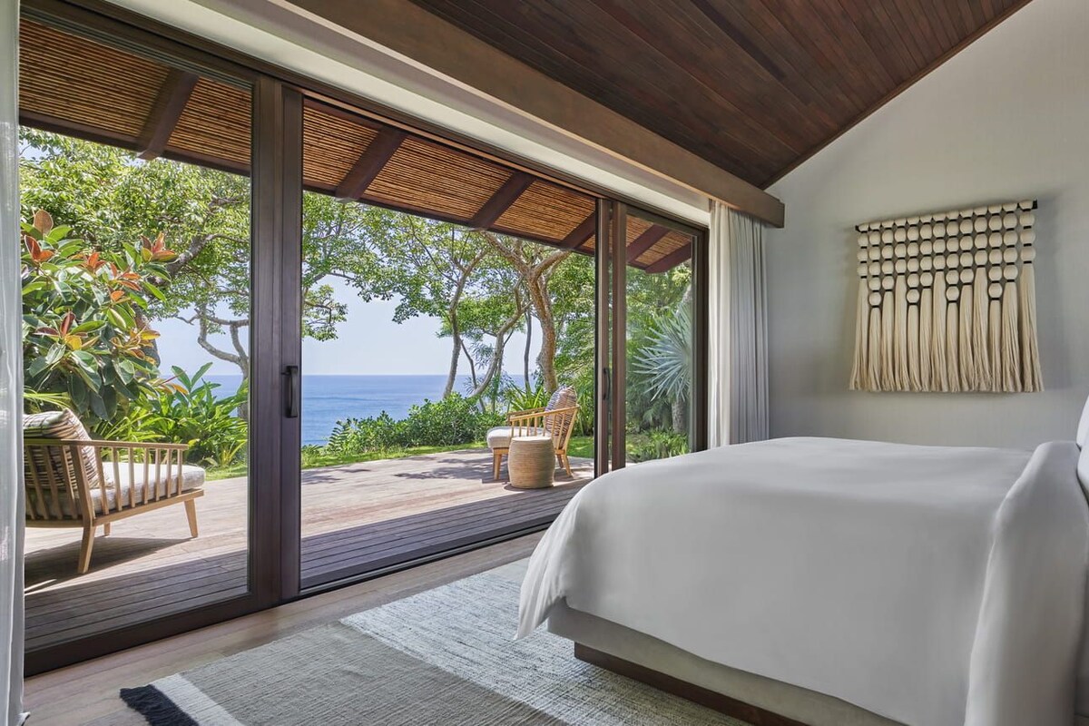 Ein Schlafzimmer mit einem großen Bett, Meerblick und einer Schiebetür zu einem Außenbereich mit Sitzgelegenheit.