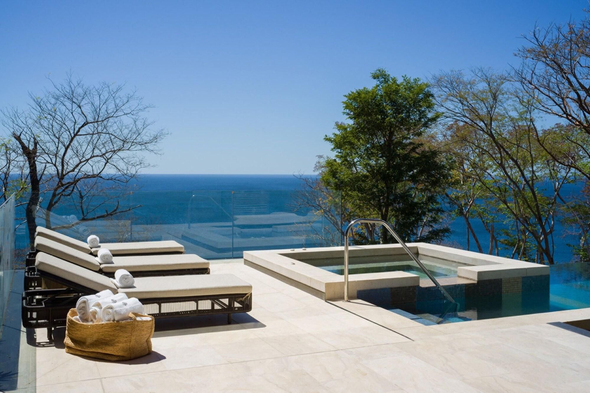 A rooftop infinity pool with a view of the ocean, lounge chairs, and a hot tub.