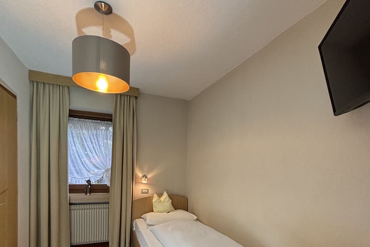 A hotel room with a single bed, a desk, a chair, a window with curtains, a radiator, and a wall-mounted TV.