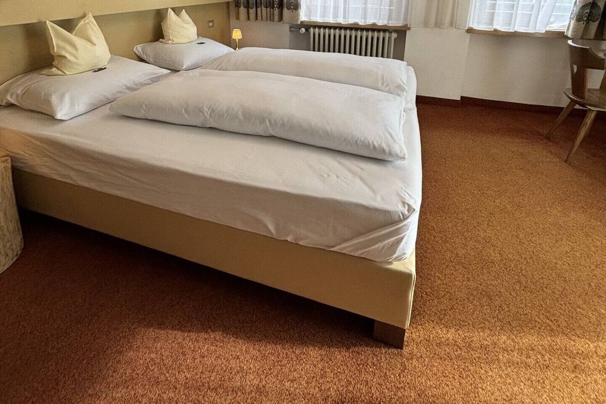 A neatly made bed with white linens, a radiator under the window, and two framed artworks on the wall.