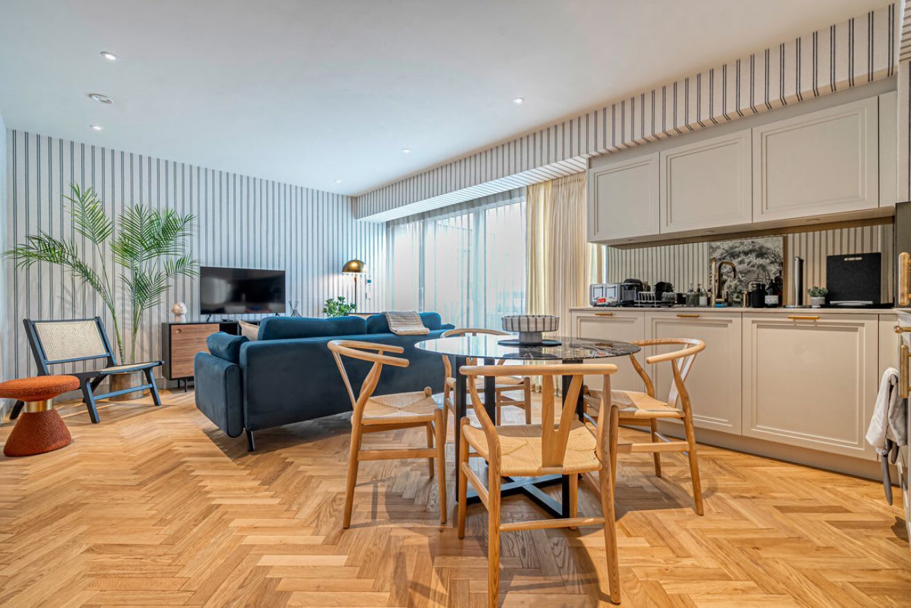 Uma sala de estar moderna com um sofá azul, cadeiras de madeira, piso em padrão herringbone, uma TV e uma área de cozinha com armários e eletrodomésticos.