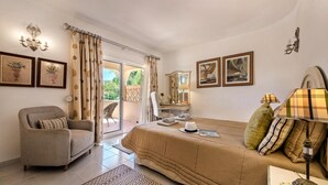 Ein helles Schlafzimmer mit einem Bett, einem Sofa, einem Sessel und einem Schreibtisch. Vorhänge, eine Chandelieber und gerahmte Bilder schmücken die Wände.