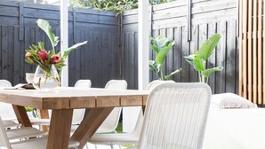 A wooden table with white chairs, a vase with flowers, and a view of a wooden fence and greenery. - Rosebud’s Modern Coastal Escape • Walk to Beach • Sleeps 10 (Rosebud)