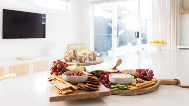A well-lit room with a large dining table set for a meal, featuring various cheeses, fruits, and crackers. - Rosebud’s Modern Coastal Escape • Walk to Beach • Sleeps 10 (Rosebud)