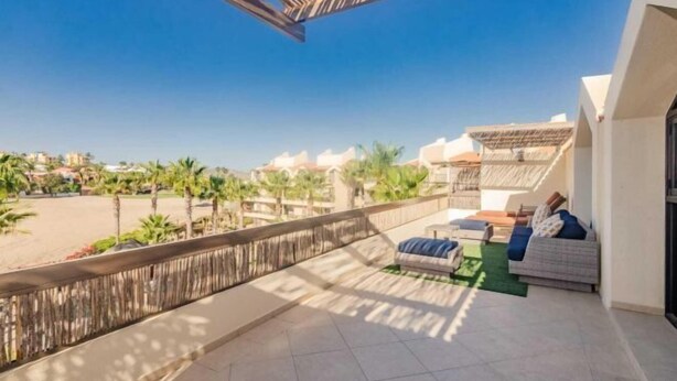 A balcony with a view of palm trees and buildings, a sofa, and a small table. - 2BD Condo in La Costa Phase 2! (San Jose del Cabo)