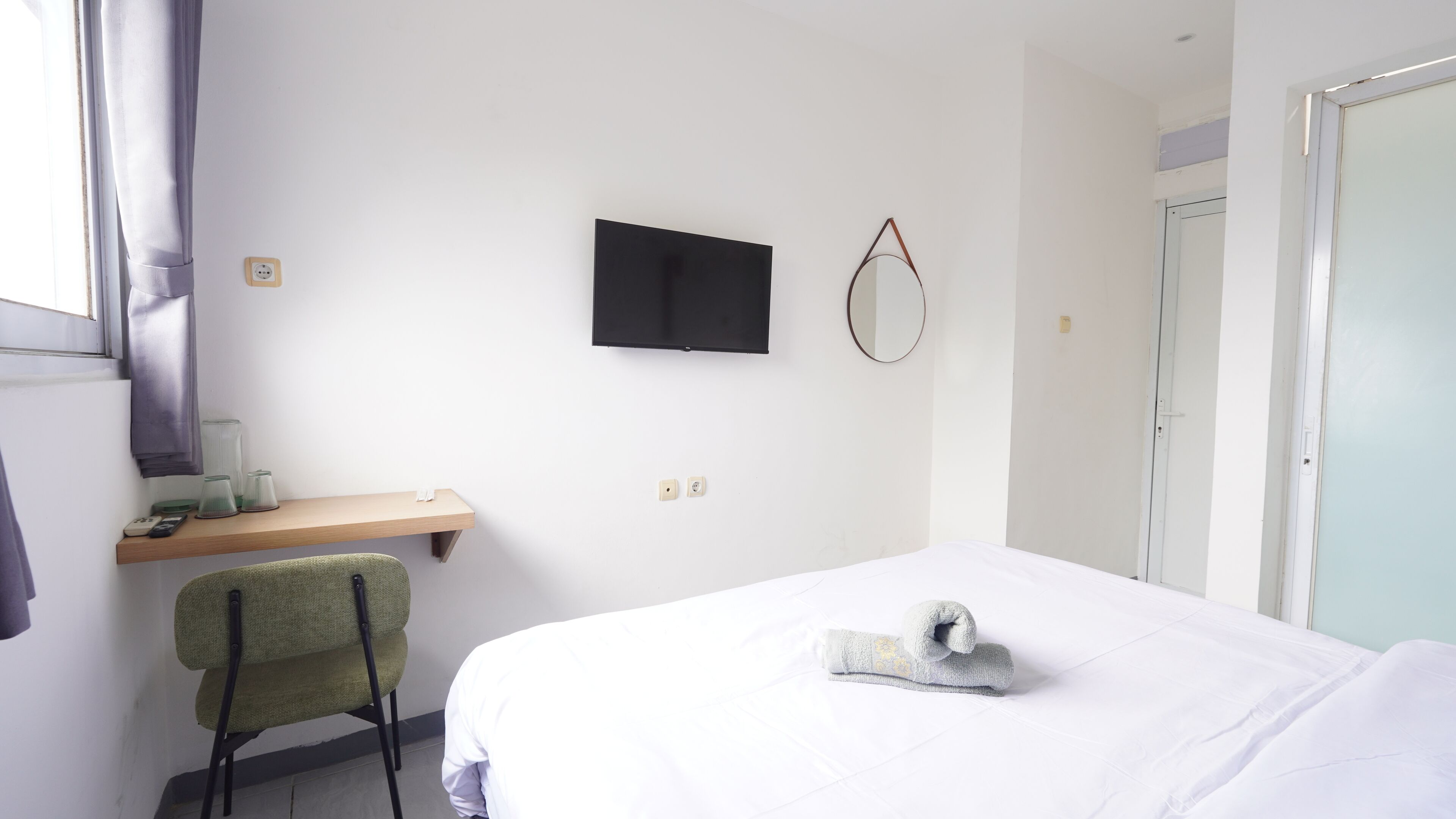 Una habitación de hotel con cama, un escritorio con silla, un televisor montado en la pared y una puerta.