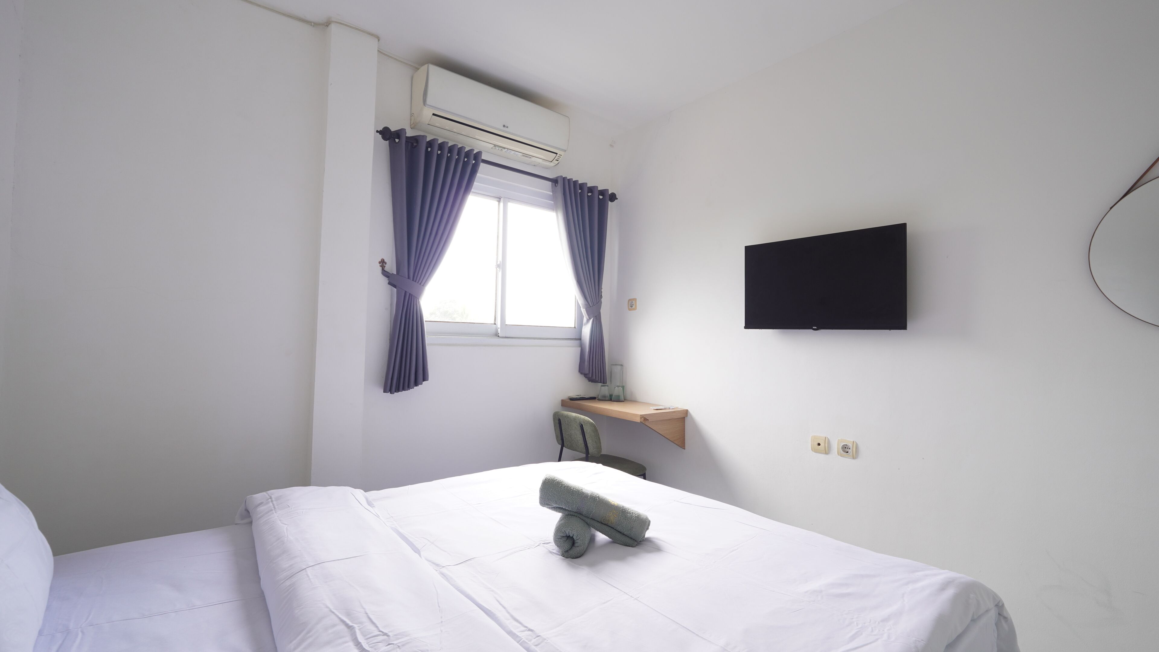 A hotel room with a bed, a desk with a chair, a flat-screen TV mounted on the wall, and an air conditioning unit above the window.