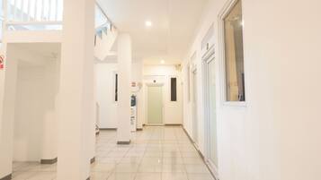 A bright, modern hallway with white walls and tiled floors. There are doors on both sides, a water dispenser, and a fire alarm.