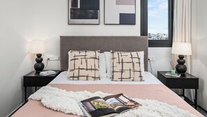 Una habitación de hotel moderna con una cama grande, dos mesitas de noche, un ventanal con vistas a la ciudad y una obra de arte abstracto en la pared.