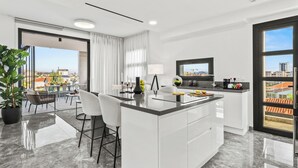 A modern kitchen with a large island, a dining area with chairs, and a view of the city through large windows. - Noir Luxury Residence - Rooftop & Jacuzzi by Nomads (Limassol)