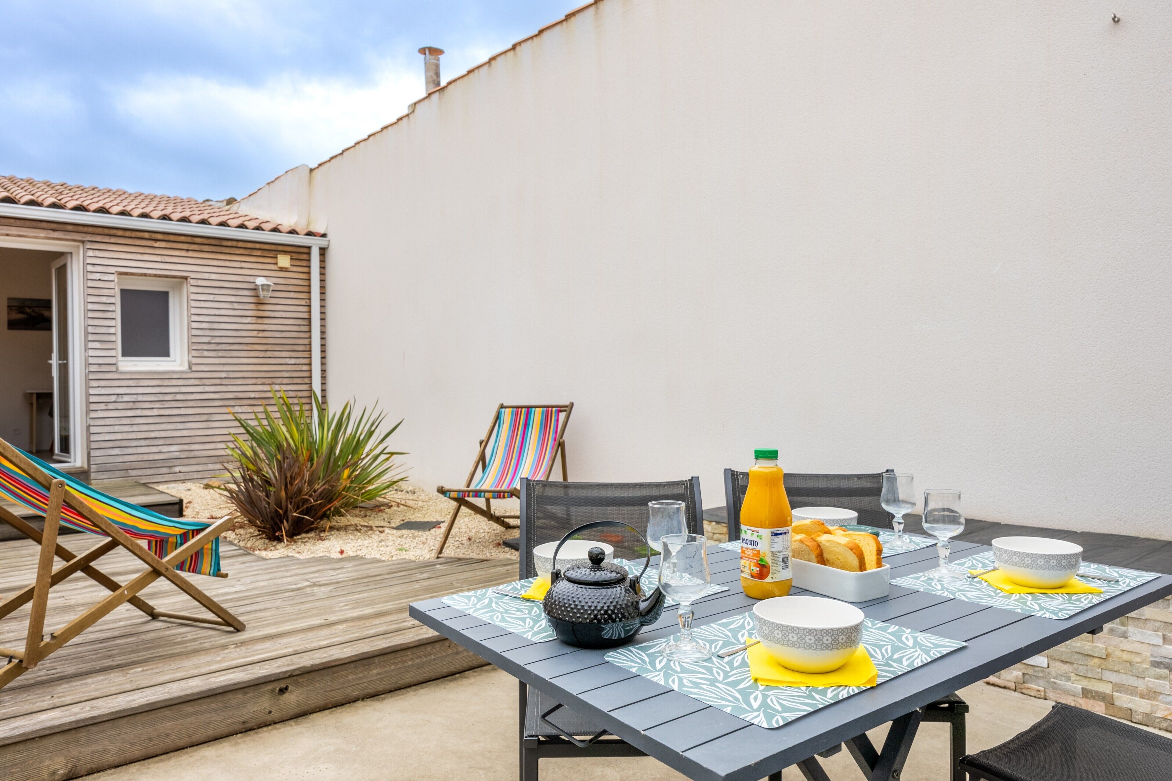 Une terrasse avec une table mise pour le petit-déjeuner, comprenant une théière, des verres et du jus d’orange.
