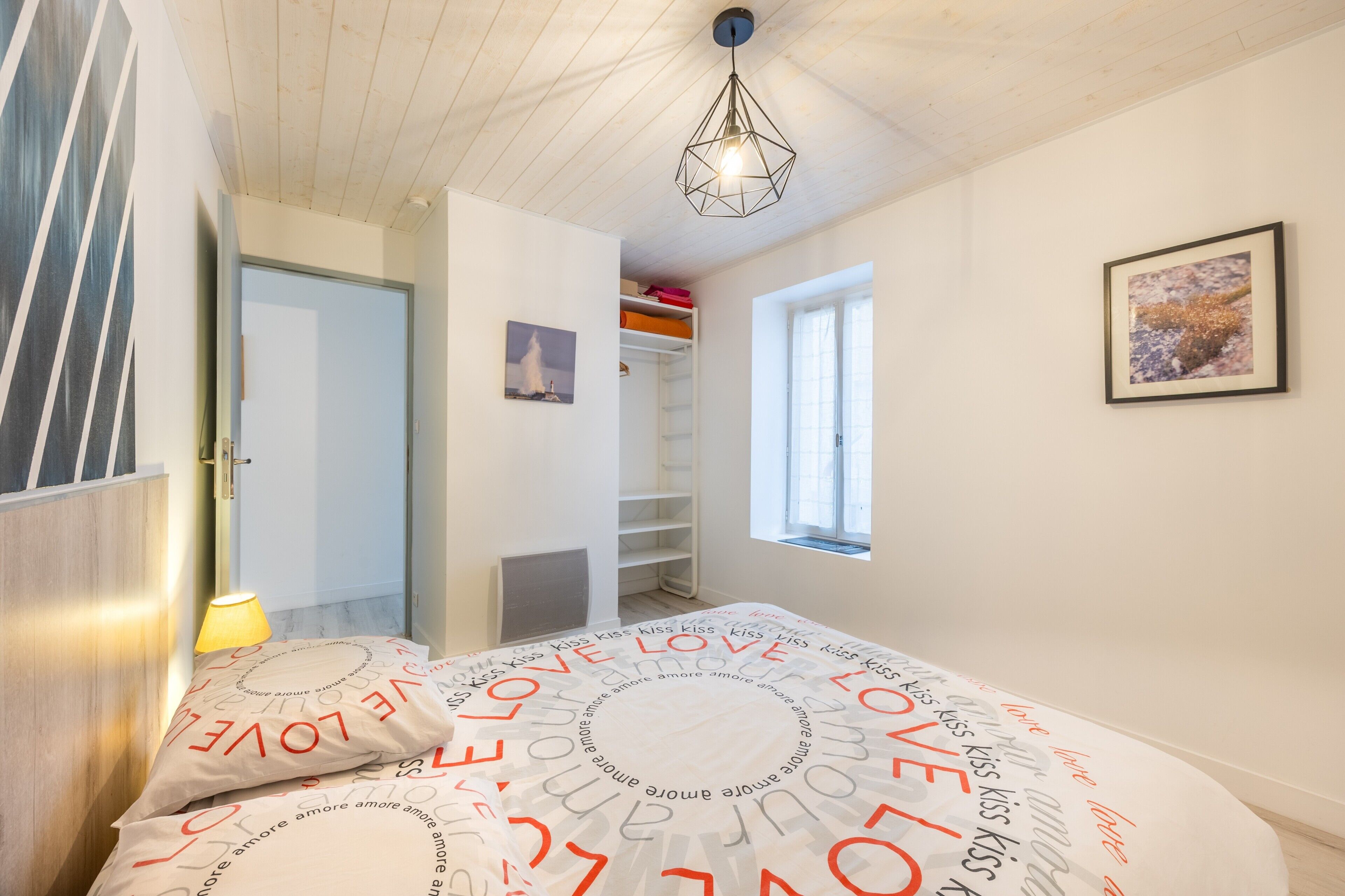 Un dormitorio con cama, lámpara de noche, armario, ventana y un cuadro en la pared.