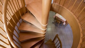 Uma escadaria em espiral com guarda-corpos e degraus de madeira, descendo para um andar inferior com uma pequena mesa e cadeiras.