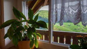 Janela com vista para uma paisagem verde, uma planta em vaso e cortina de crochê.