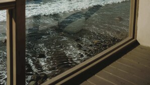 Une vue à travers la fenêtre d’un balcon en bois donnant sur la mer, où les vagues s’écrasent sur le rivage.
