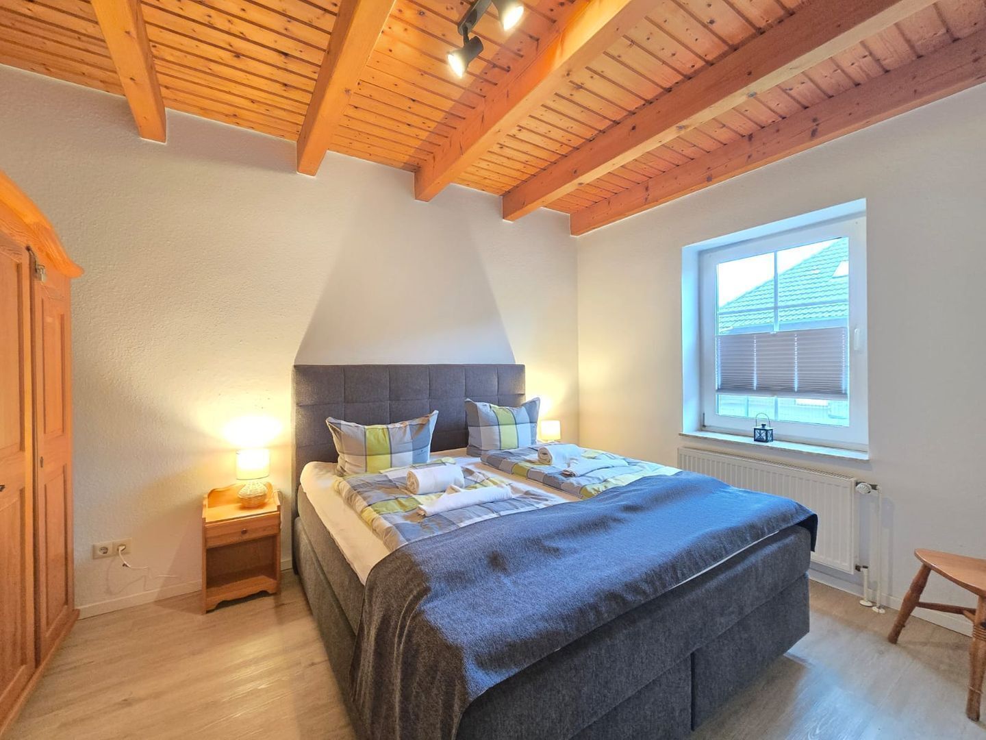 Ein Schlafzimmer mit einem Bett, Holzbalkendecke, einem Fenster mit Jalousien und einem Nachttisch mit Lampe.