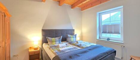 Ein Schlafzimmer mit einem Bett, Holzbalkendecke, einem Fenster mit Jalousien und einem Nachttisch mit Lampe.