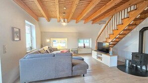A spacious living room with a grey sectional sofa, wooden ceiling beams, a TV, and a black wood-burning stove. - Ferienhaus Strandkrabbe - Comfortable vacation home Strandkrabbe with large garden (Neßmersiel)