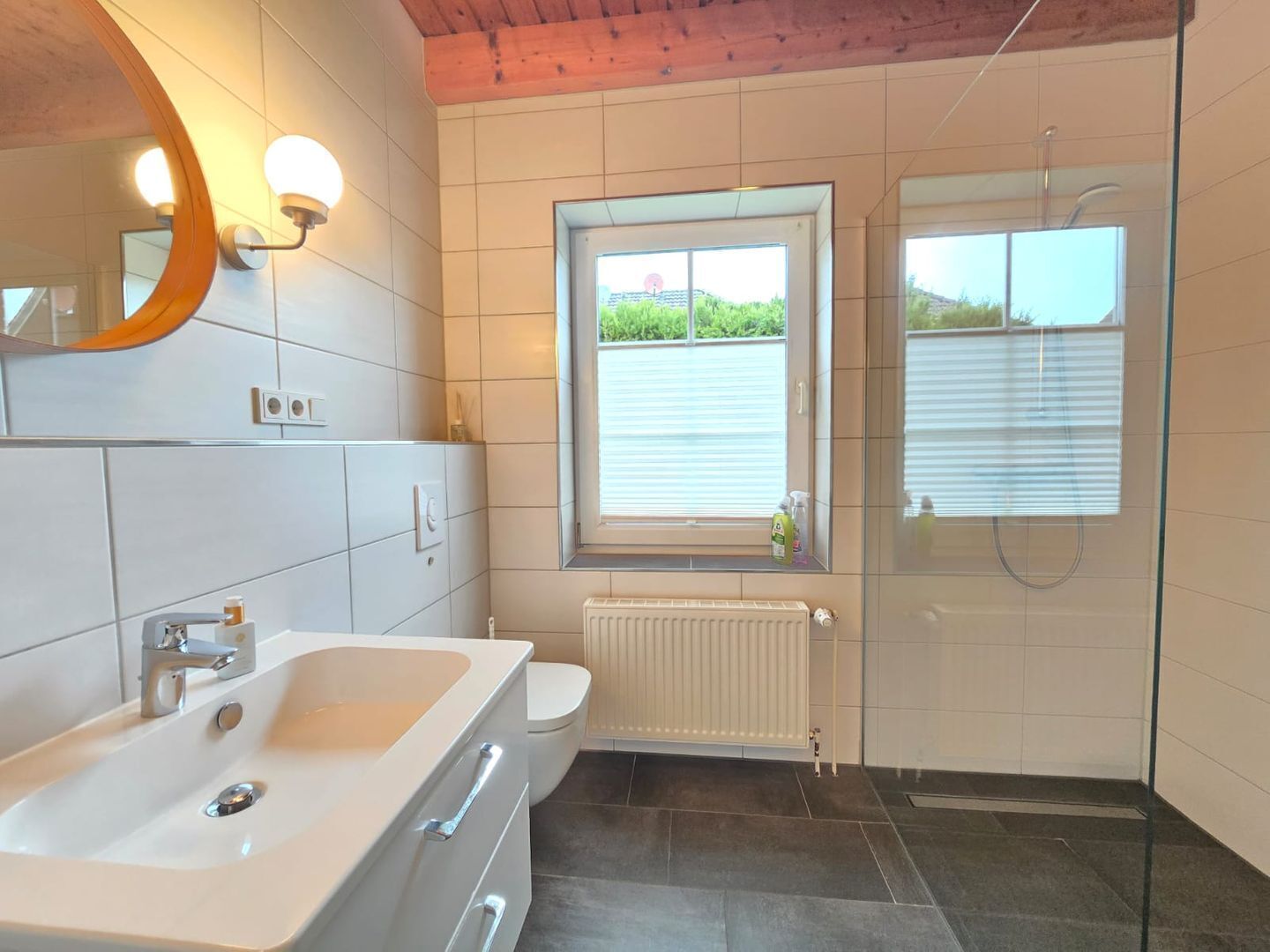 A modern bathroom with a large white bathtub, a toilet, a glass shower enclosure, and a window providing a view of greenery outside.