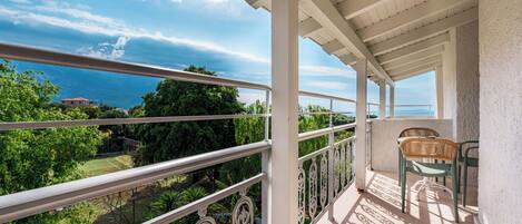 A balcony with a view of the sea, a tennis court, and green chairs.