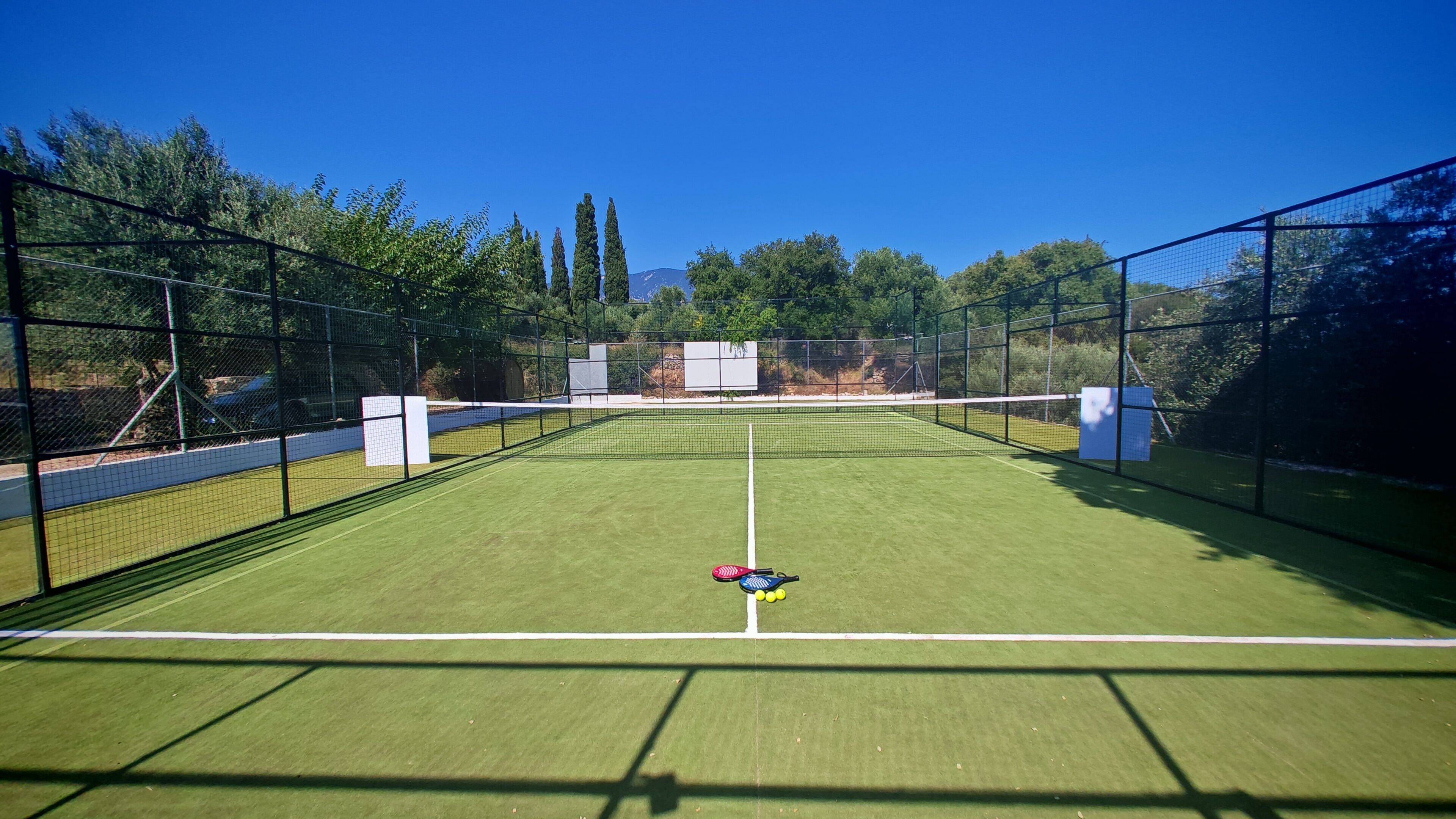 An outdoor tennis court with a net, surrounded by trees and a clear sky.