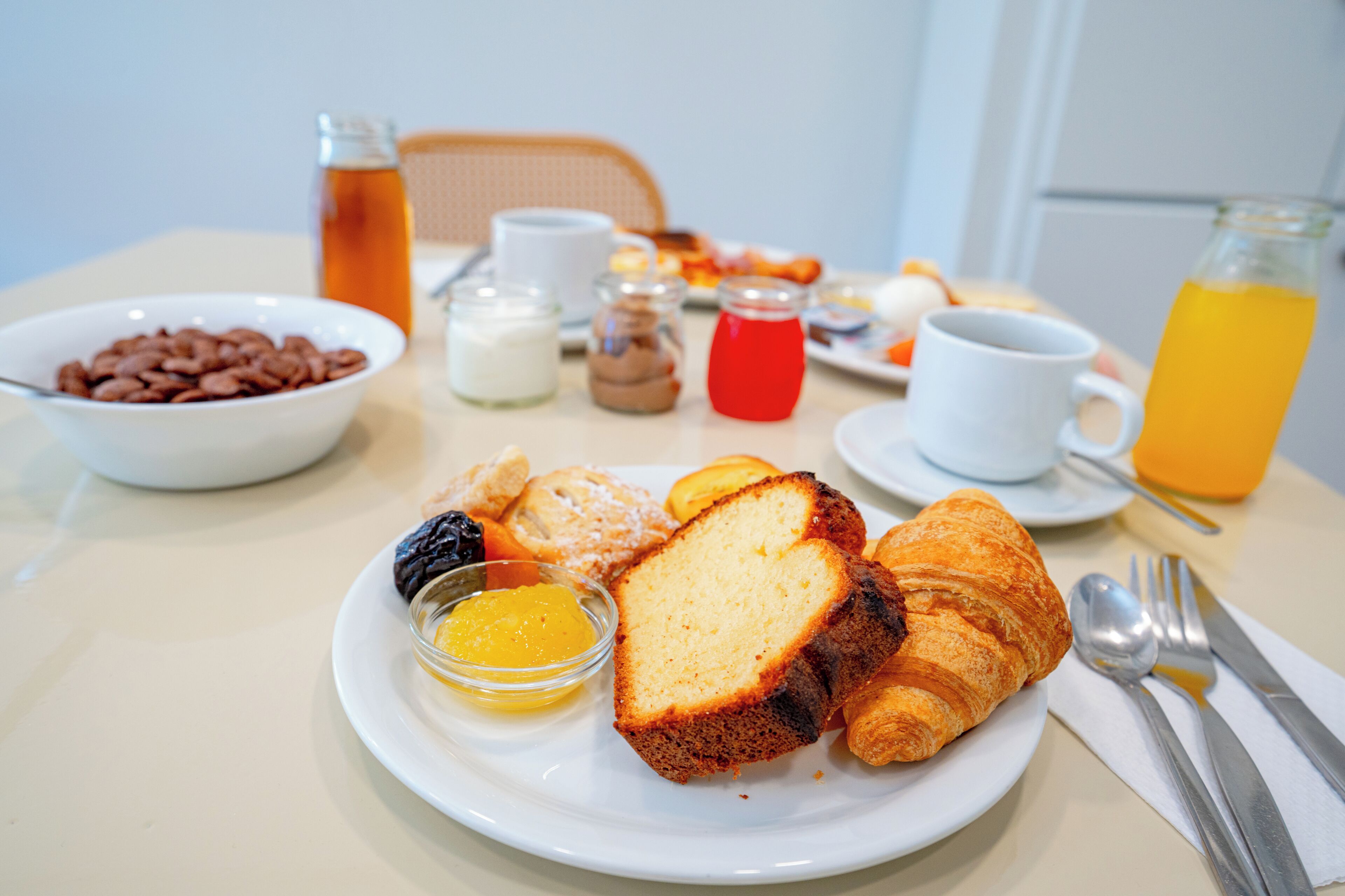 A breakfast setting with croissants, a bowl of cereal, a glass of orange juice, and a bottle of tea.