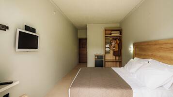 Habitación de hotel con cama, televisor montado en la pared, armario con ropa colgada y un pequeño escritorio con lámpara.