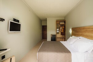 Habitación de hotel con cama, televisor montado en la pared, armario con ropa colgada y un pequeño escritorio con lámpara.